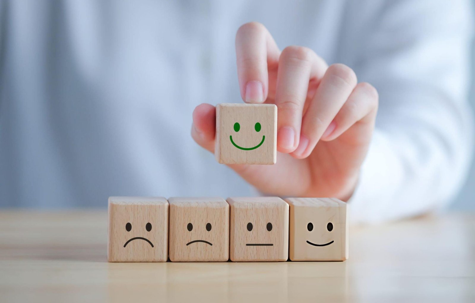 "A hand holding a wooden block with a green smiley face, above four other blocks with various facial expressions (sad, neutral, happy). This image represents different emotional states and suggests choices for positive mental health outcomes. Relevant to Mental Health Therapy Options.