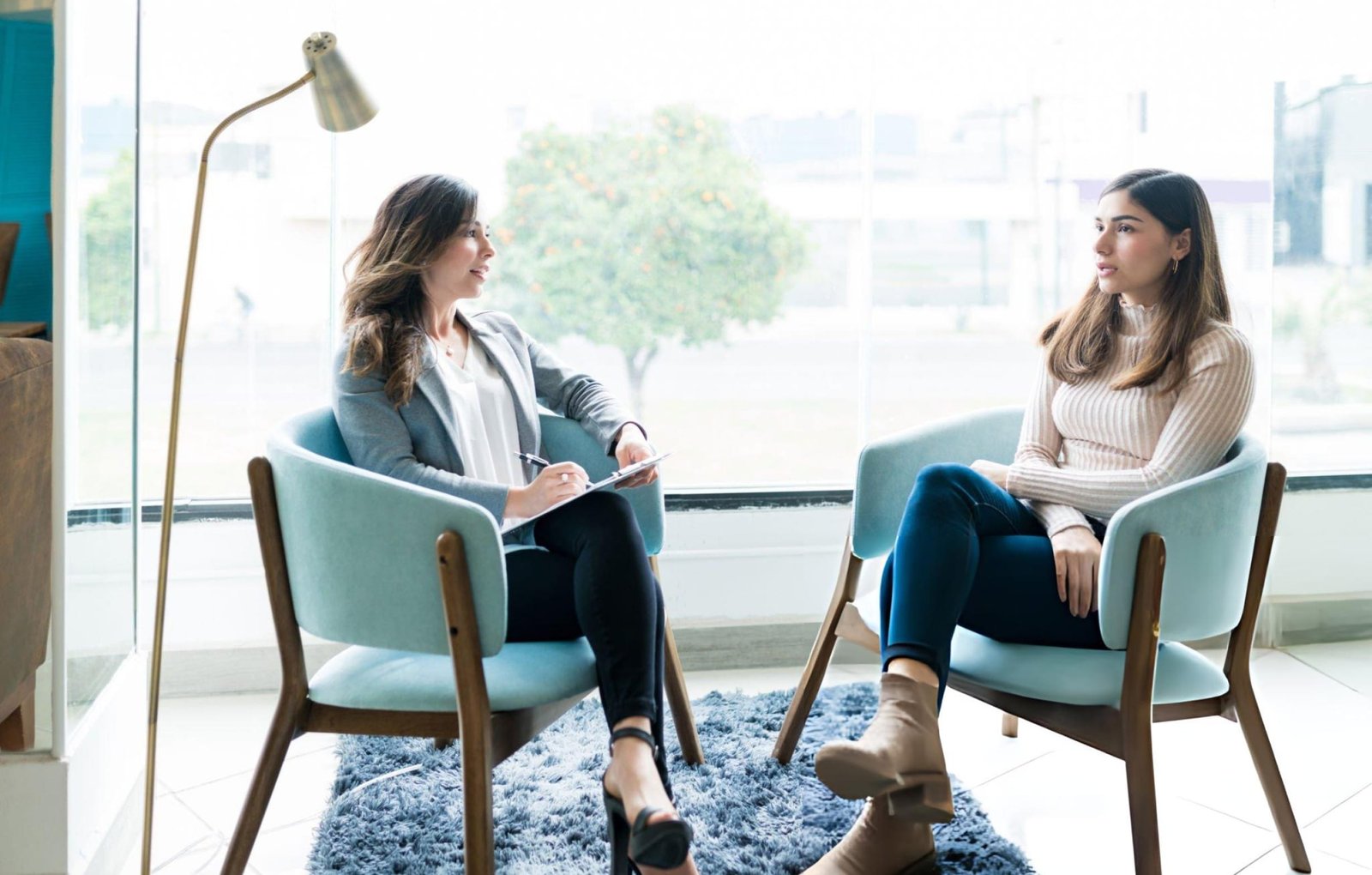 Two individuals seated in light blue chairs face each other in a professional setting, indicative of a therapy session. One holds a clipboard and pen, clearly a therapist, while the other sits with legs crossed. The setting includes a floor lamp, a blue rug, and a large window with a view of trees and buildings. This image represents DBT for anxiety.