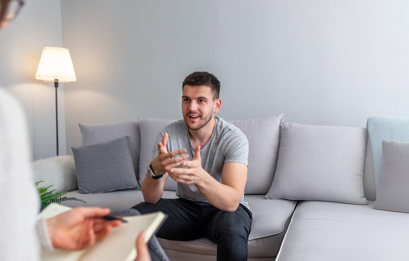 "A person sitting on a couch, engaged in conversation with a therapist who holds a notebook and pen. The setting is a comfortable, private room with soft lighting and cushions. This image represents individual therapy.